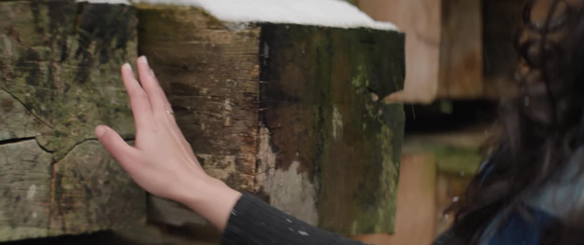 Woman touches ends of large beams of weathered lumber as snow falls