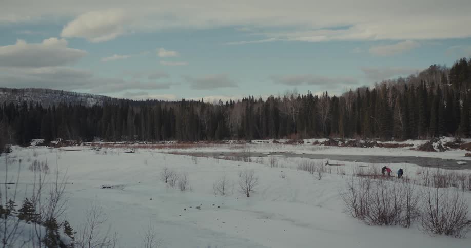 People stand on icy shore across narrow strip of water from conifer forest on a winter day