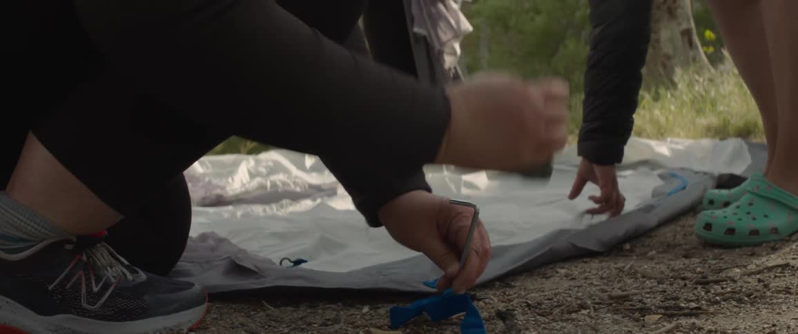 Three people work to put up a tent in a forest clearing with one hammering a tent pole on the ground