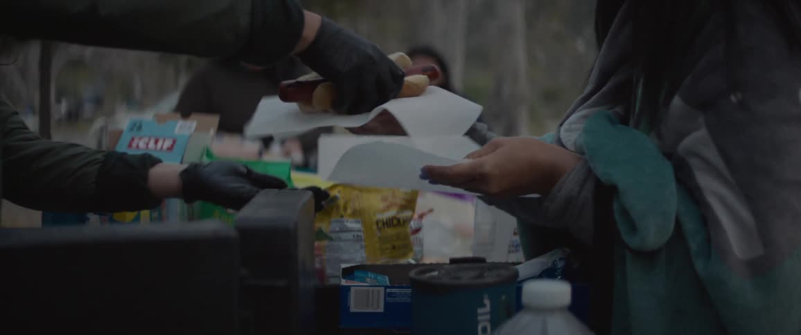 A group of woman are around a picnic table filled with camp food outdoors in a wooded area with one handing hotdogs to another on a cool evening