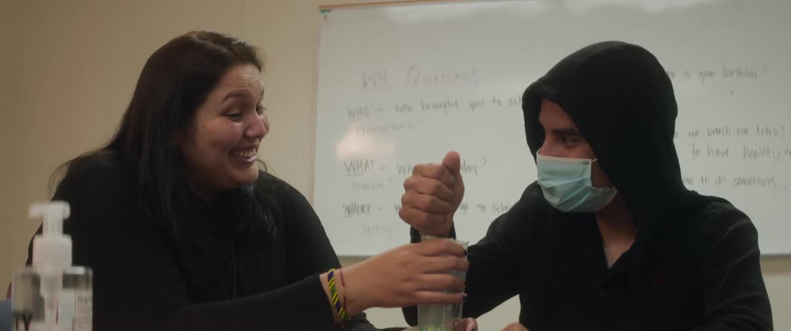 Two people sitting at a table in an indoor room with one of them pouring colorful granules into a cup
