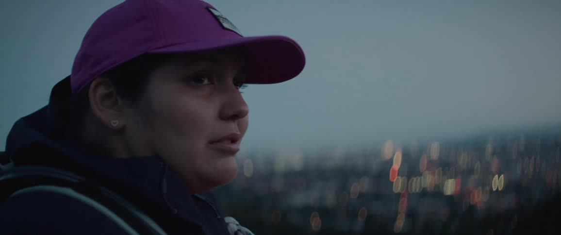 Woman wearing a baseball cap looks out over blurred twinkling lights in the background with a sad expression of holding back tears in the late evening