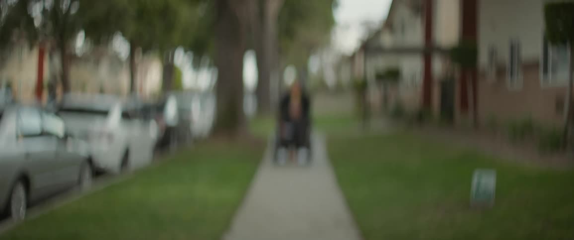 Out of focus view of a person pushing another person down the sidewalk between a line of cars parked on the curb and townhome apartments during the day