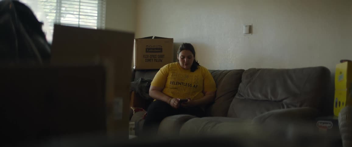 A young woman is sitting on a couch texting in a room with moving boxes everywhere and natural light coming through the window