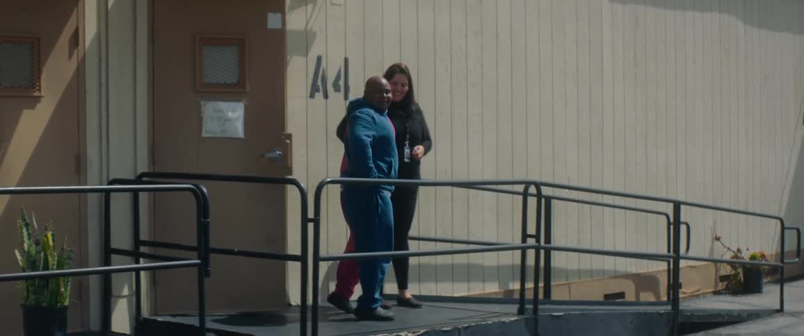 A woman walks a bald male down a ramp leading outside of a building