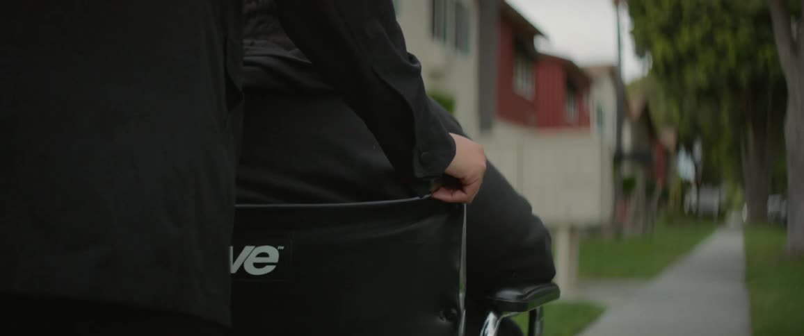 Two people in black jackets one pushing a wheelchair and the other riding move down a sidewalk lined with apartments before sundown