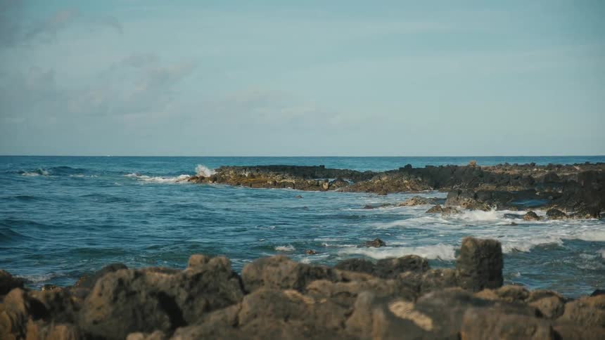 Ocean waves crashing against rocks on a beach on a day with a clear blue sky and few clouds