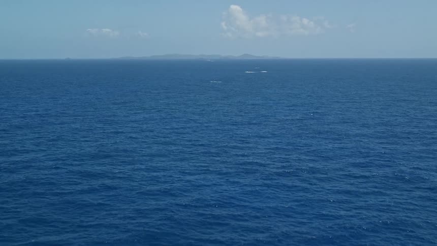An ocean with a very blue surface and a huge rock sticking out and on the horizon there are mountains