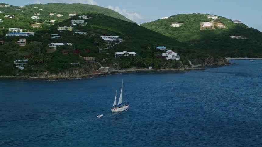 Aerial view of a sailboat passing next to a rural area on a cliff surrounded by vegetation during day time