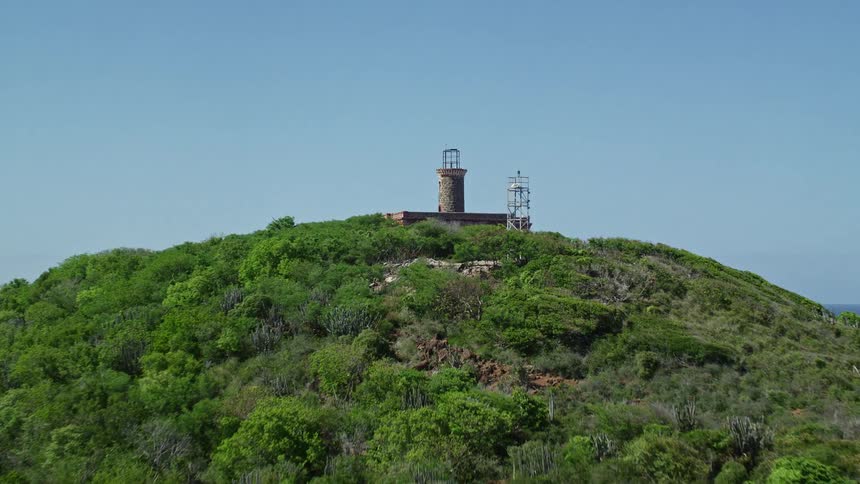 An island with a forest of trees and at the top an abandoned building and around this island there is a large ocean and some boats parked on the coast