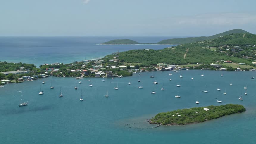 Several boats parked in the waters of a bay next to an island with residences and on the other side is the open sea