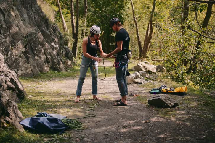 Man and woman wearing helmets outdoors on a pathway in the middle of a forest tying hiking ropes to each others waists during day time