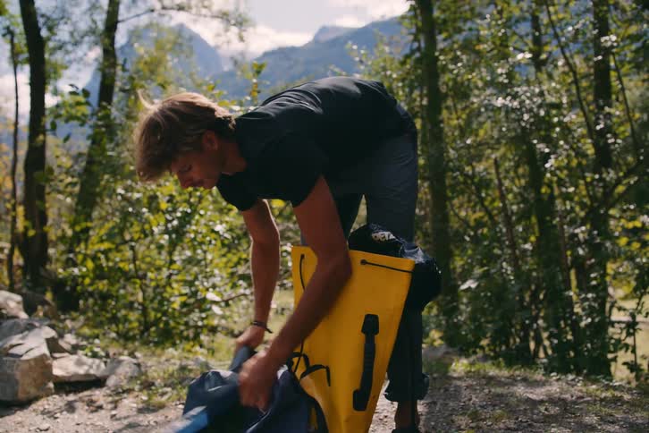 A man is removing objects from a backpack and he is in an environment with many trees