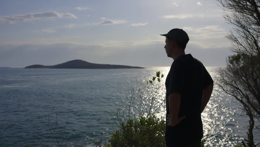 A man with his hands on his waist observing the view of the sea with sunlight reflection from a hill