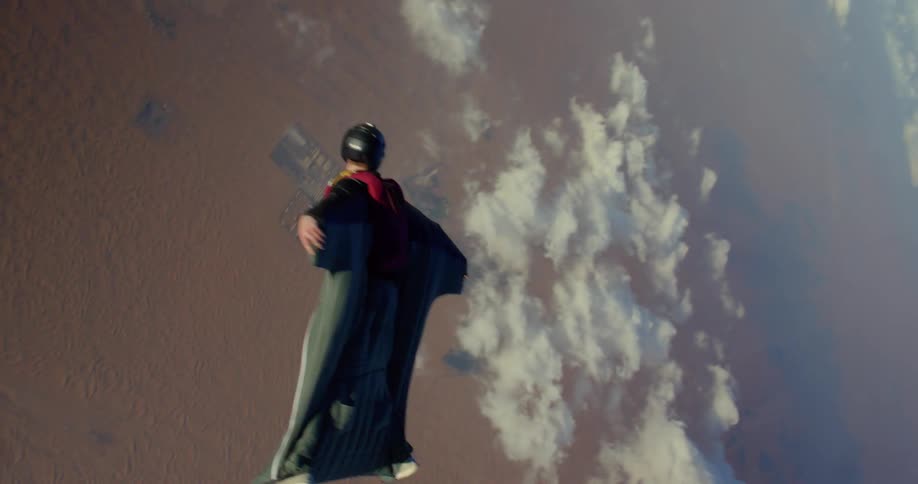 People skydive in wingsuits over desert terrain on a cloudy day
