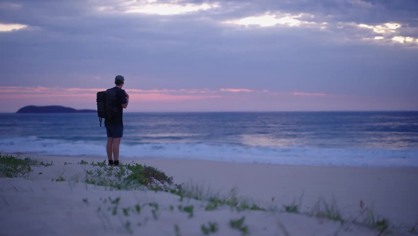 A man standing in front of the sea watching the sun set on the horizon