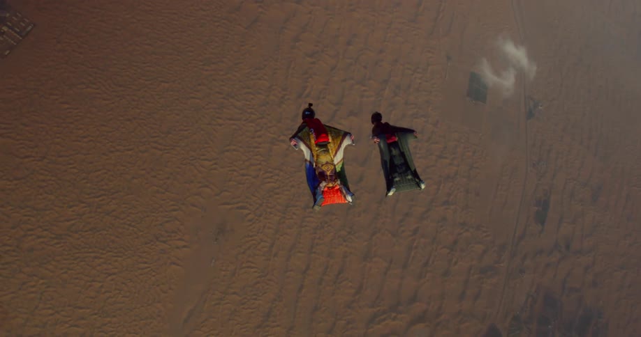 Two people are stopping while wearing wingsuits and on the ground there is a desert region