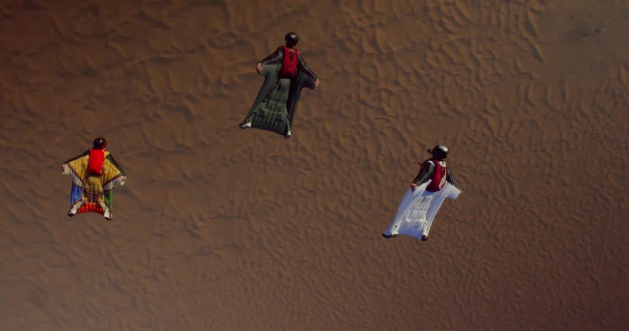 Three people are gliding while doing wing suit towards the ground in a desert region