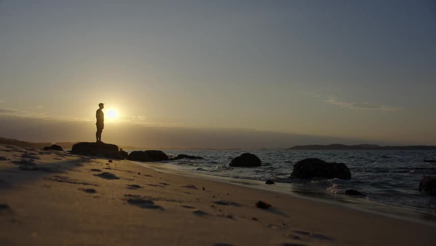 Person stands on rock on sandy ocean shore sun over horizon