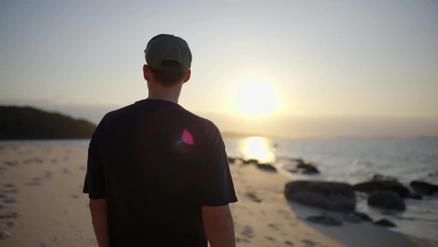 A man with his back is walking on a beach with sunlight reflecting off the sea