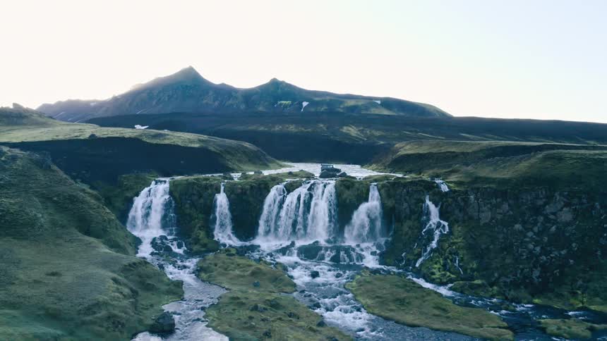 Vehicle fords river close to waterfalls with mountain in the background