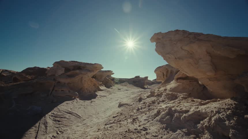 A sun shining in the blue sky in a region with some rocks and a sandy path in the middle of them