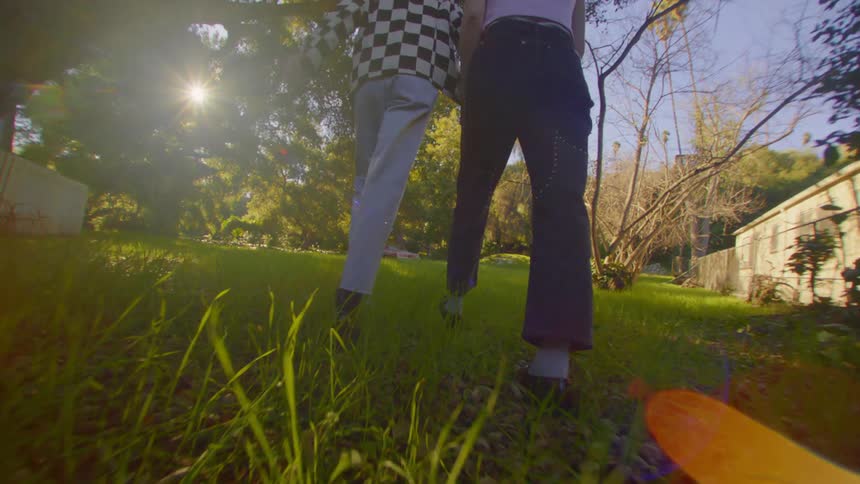 Two people wearing pants and footwear walk holding hands across a green lawn