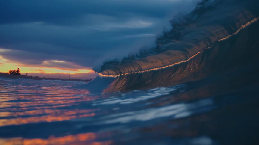 View of an expansive wave breaking in the sea against a reddish dawn sky with morning clouds