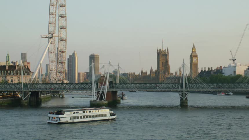A boat sailing at sunset under a bridge on the Thames in London with London Eye and Big Ben in the background