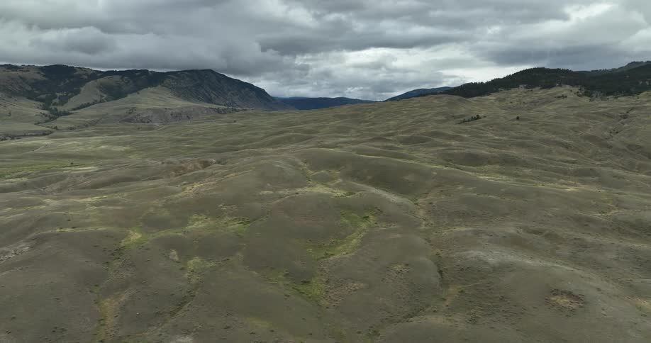 A region with a green field with many slopes and in the background there are some mountains and in the sky many dark clouds