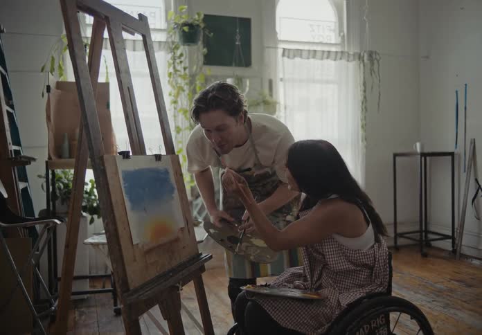 Two painters a man standing and a woman on a wheelchair look at the womans painting both laughing and playing while he gives suggestions