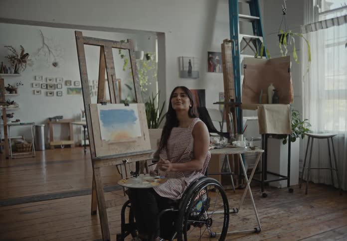 Woman in a wheelchair holding a paint palette with a brush and mixing the colors with a slight smile