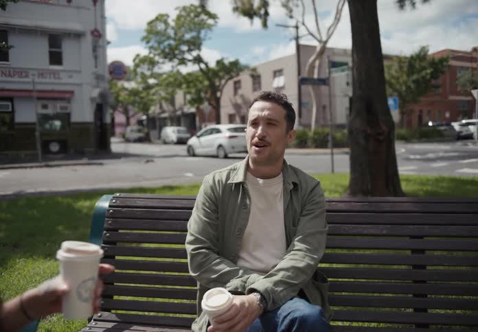 Man and woman sit in park talking as car passes through intersection in the background