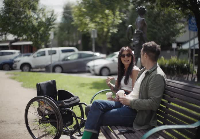 A man and a woman sitting on a park bench talking with a wheelchair in front of the woman
