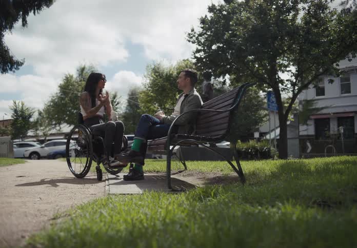 A woman in a wheelchair and a man in a park bench talk to each other