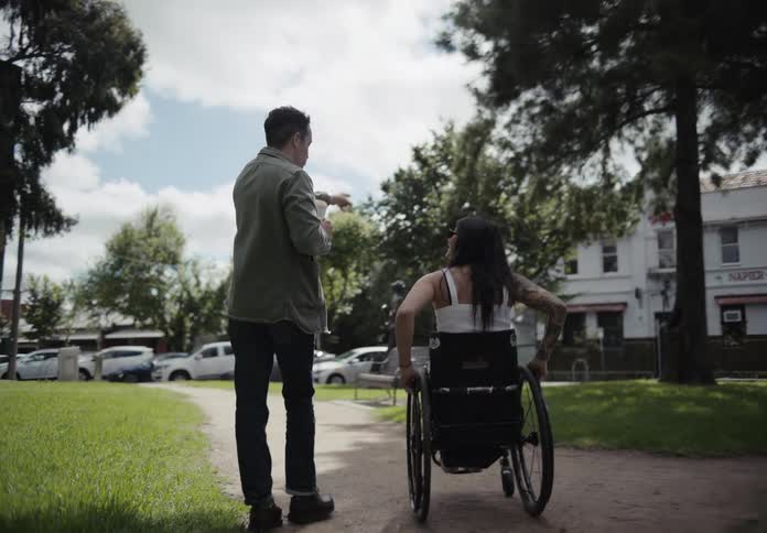 Man walking alongside a woman in a wheelchair on an outdoor sidewalk