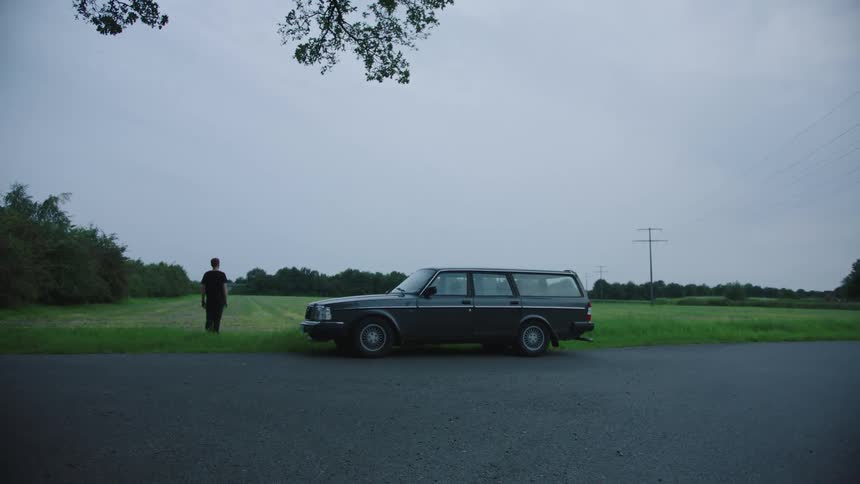 A man stands still in a grassy field next to sedan on a cloudy summer day