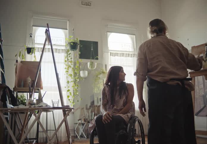 A woman in a wheelchair is talking to a woman who is standing and is pointing to a blackboard with an artistic painting