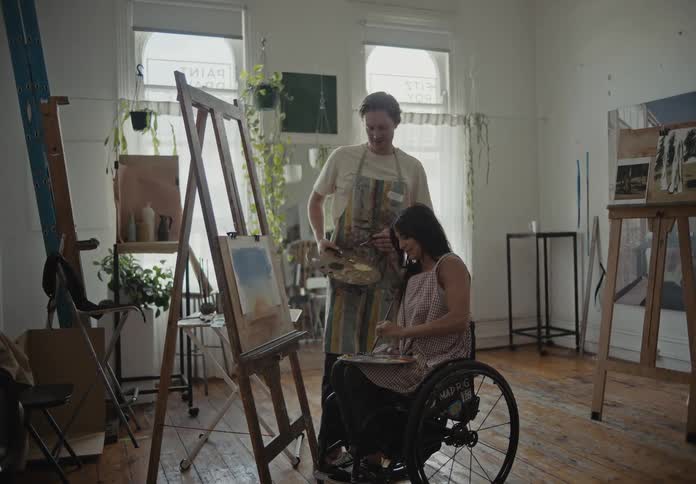 A painting session in a bright studio with a person in a wheelchair and another person on an easel