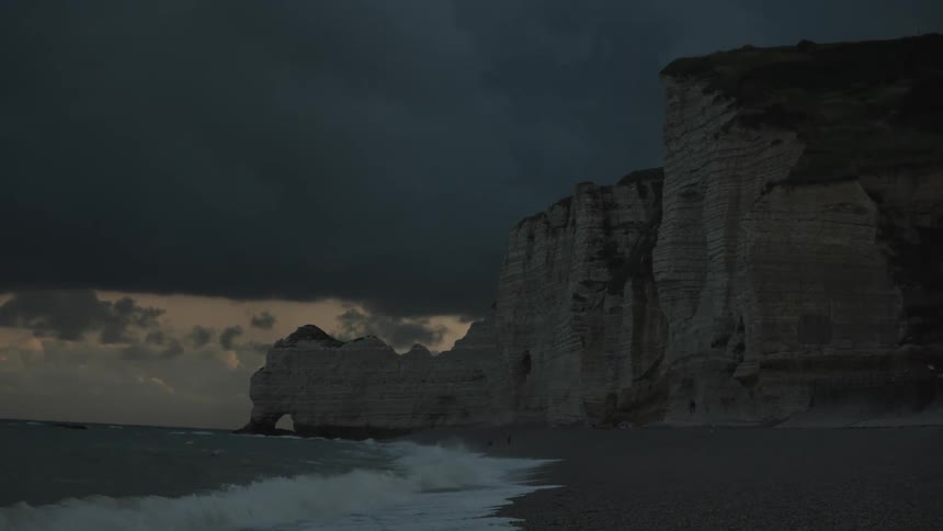 On a cloudy evening on the beach waves hit the shore as a huge rock cliff is seen