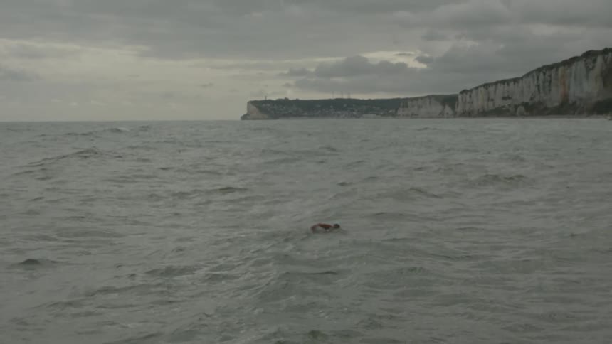 A person is swimming in a sea with several waves and close to a coast with a wall of rocks