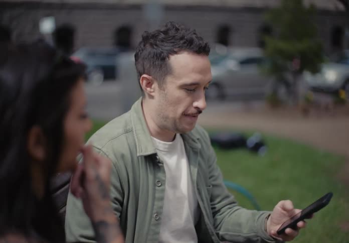 A man with his cell phone in his hand is talking and turns his head to his right to talk to a woman