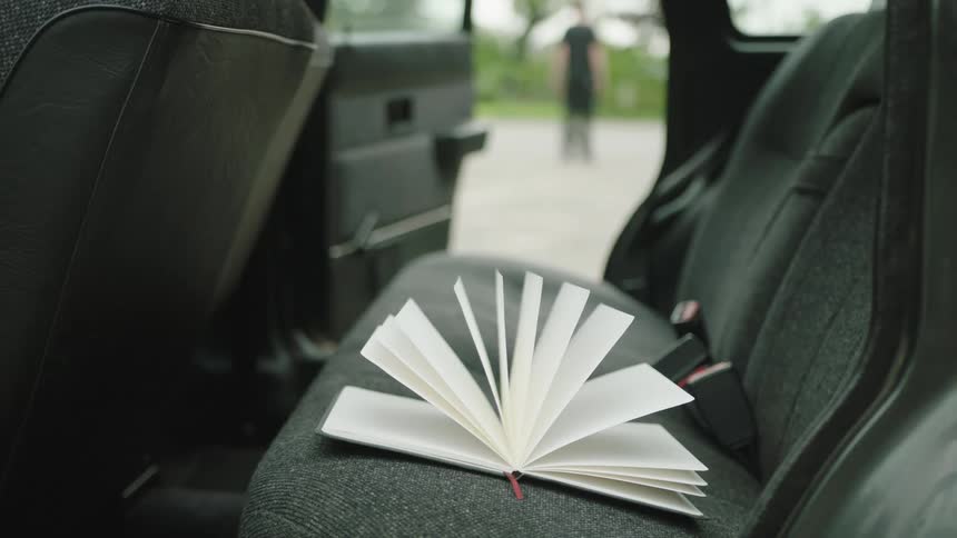 A book with white pages laying on a dark car seat and a person in the background