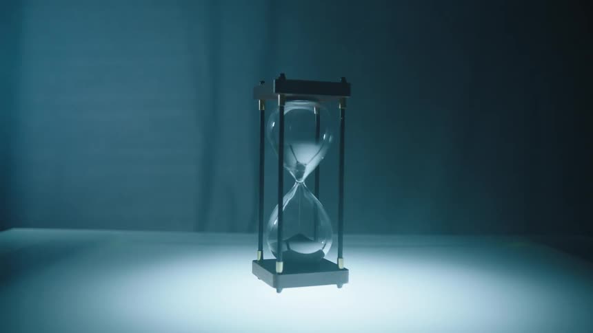 An hourglass with grains of sand inside that are falling to the bottom and the floor is illuminated