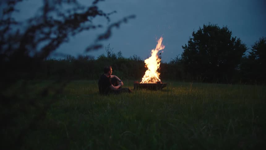 A man sitting in the countryside looking at a campfire