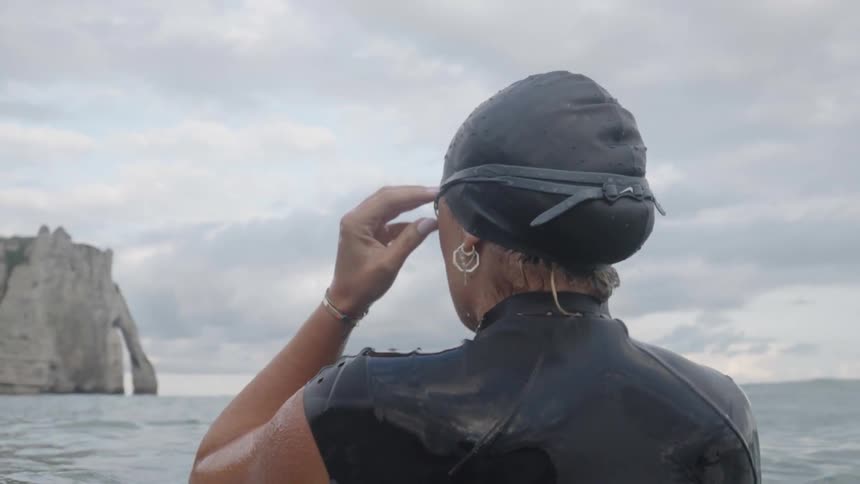 A person is standing in water adjusting a cap with rocky cliffs in the background