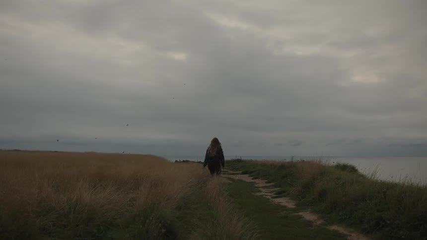Woman parallels path near coast while walking through field on an overcast day