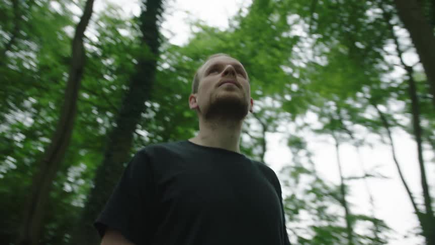 A man is in the middle of a forest with trees with green leaves and is looking to the sides