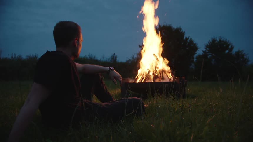 A person sitting on the grass watching a fire in a metal fire pit