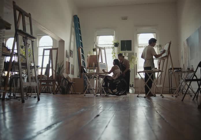 A woman on a wheelchair is painting wih a guy beside her and another guy painting behind them